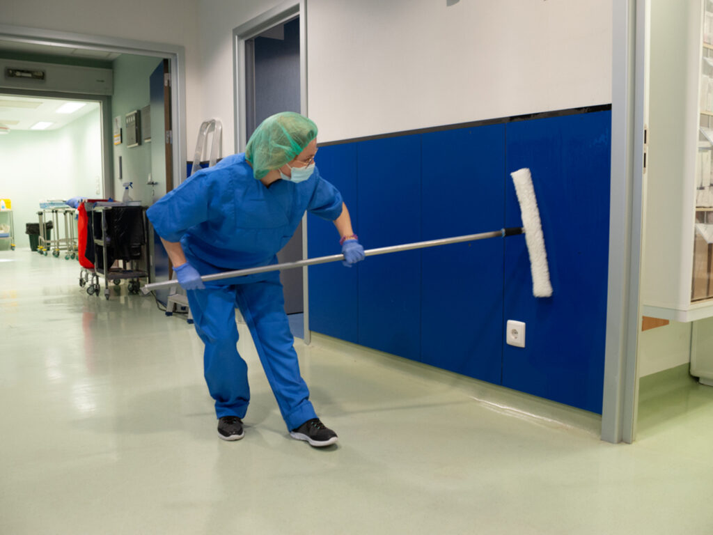 Healthcare worker cleaning hospital wall with a mop in a corridor.
