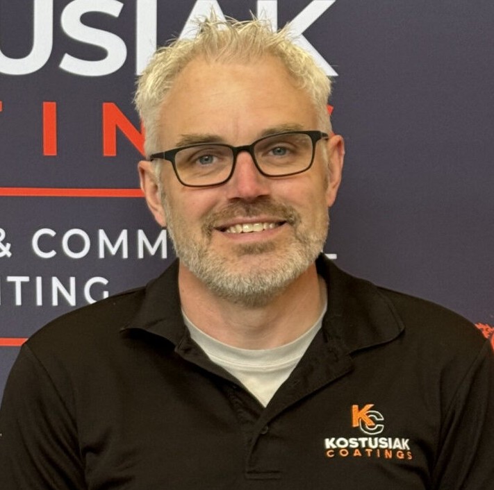 Man in Kostusiak Coatings shirt smiling in front of branded backdrop